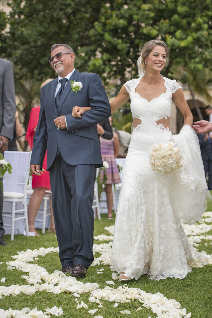 La novia entra tomada del brazo de su padre por el camino de pétalos en los jardines del hotel.