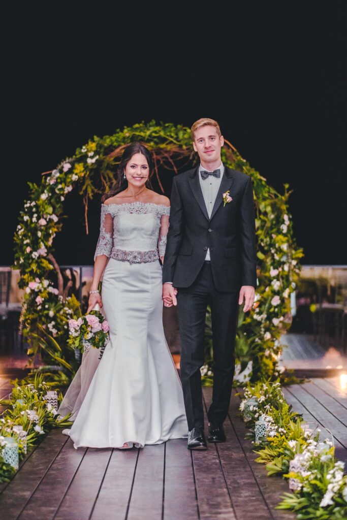 Novios caminando de la mano bajo arco floral en el Hotel Packard