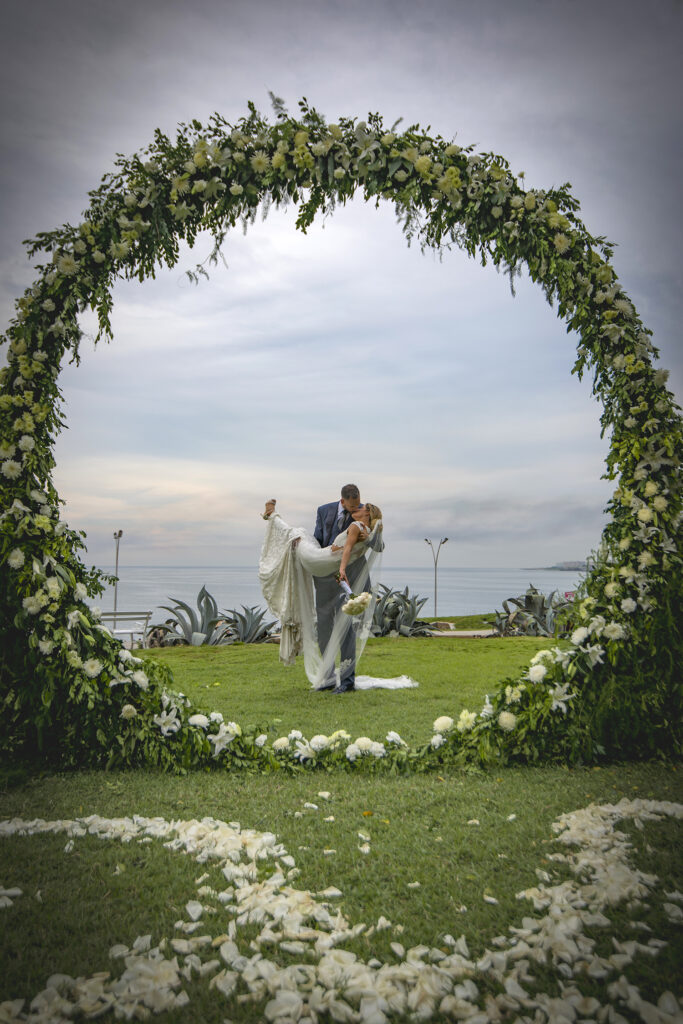 El novio carga a la novia bajo el aro floral frente al mar durante su boda en el Hotel Nacional de Cuba.