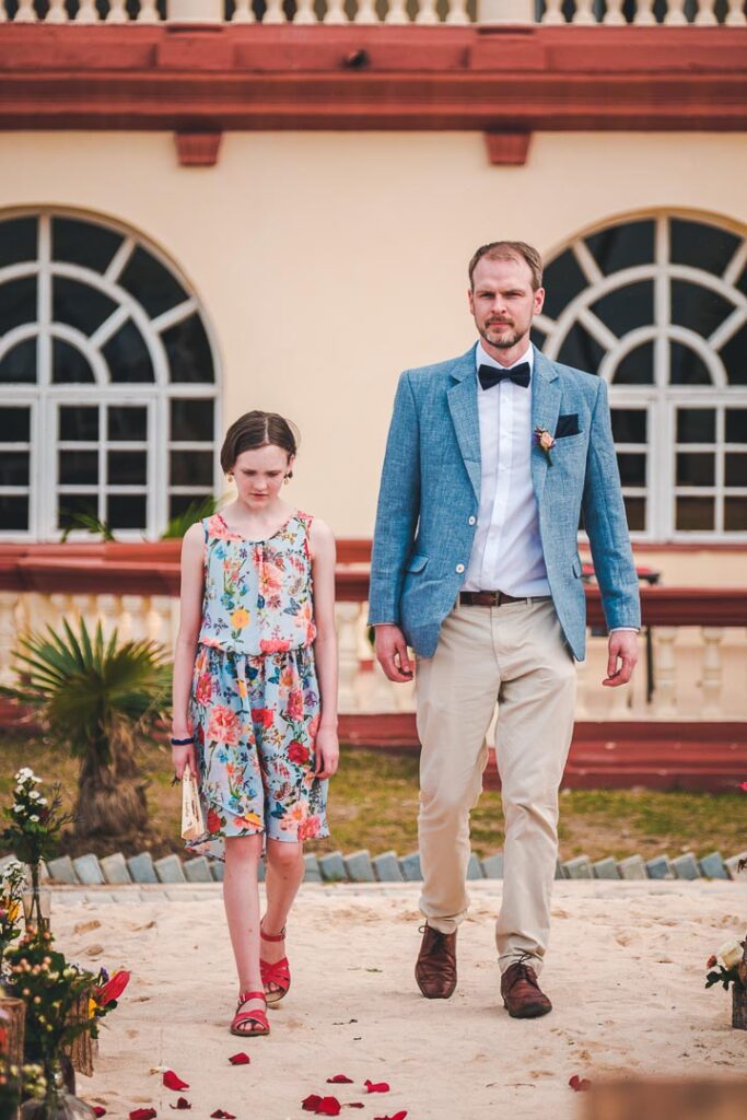 Un integrante de la corte entra a la ceremonia con una niña por el pasillo de pétalos