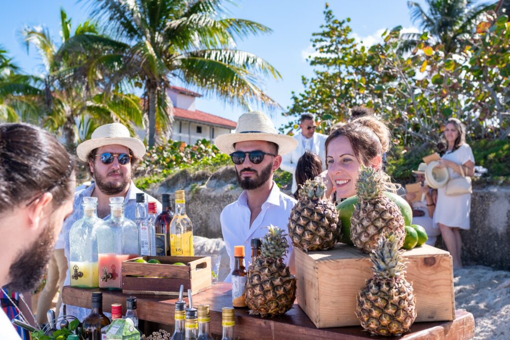 Primer plano del bar en la playa, con invitados usando sombreros y disfrutando de cócteles tropicales servidos en piñas, un ambiente perfecto para esta boda de destino en Varadero.