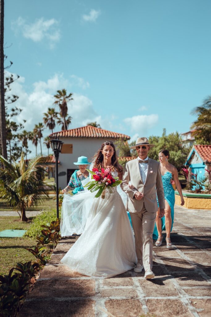 Caminando hacia la ceremonia con el brazo de su padre, mientras su madre y hermana ajustan el vestido de novia, reflejando la emoción de este momento especial.