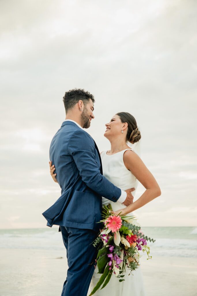 Alba y Renato mirándose tras la ceremonia, con un bouquet tropical en la playa.