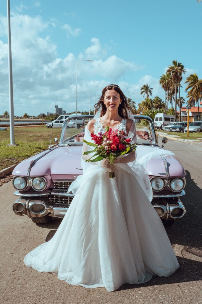 La novia posando con su ramo de novia frente al carro rosa de época, destacando los tonos vibrantes del bouquet.