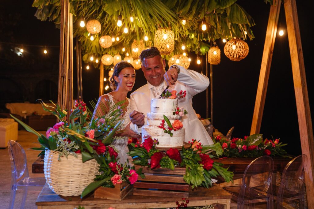 Giada y Gorke celebrando emocionados durante el corte del pastel de su boda, detalles de la decoración realzada por la iluminación al anochecer.