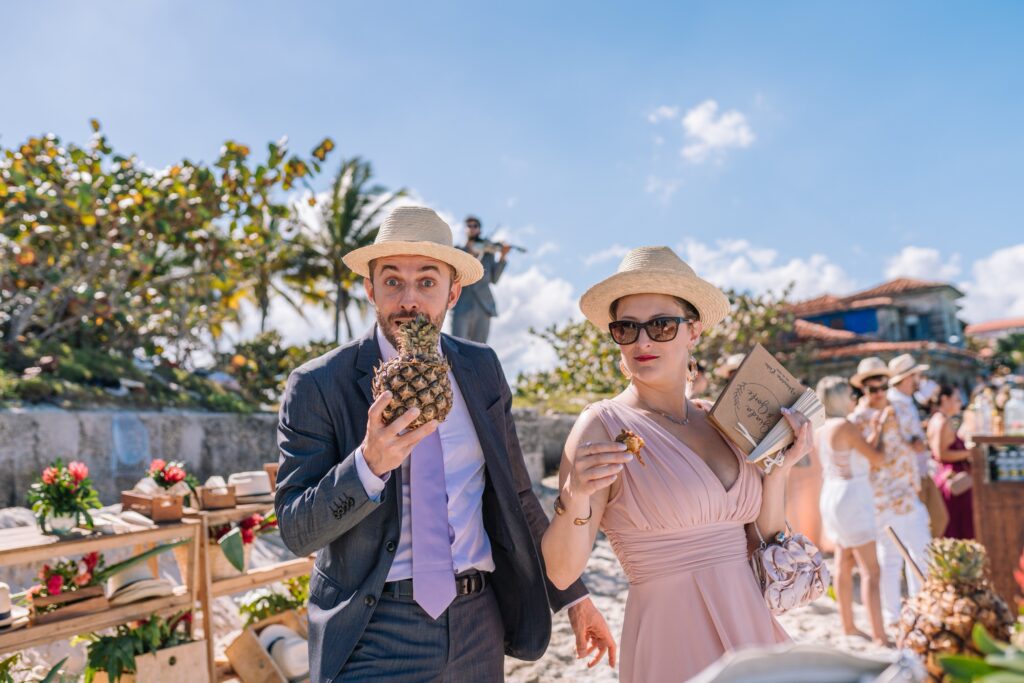 Invitados pasando mientras disfrutan de una piña colada servida en la piña, con la estación de regalos decorada con flores, sombreros y abanicos al fondo, en la boda de destino en Varadero.”