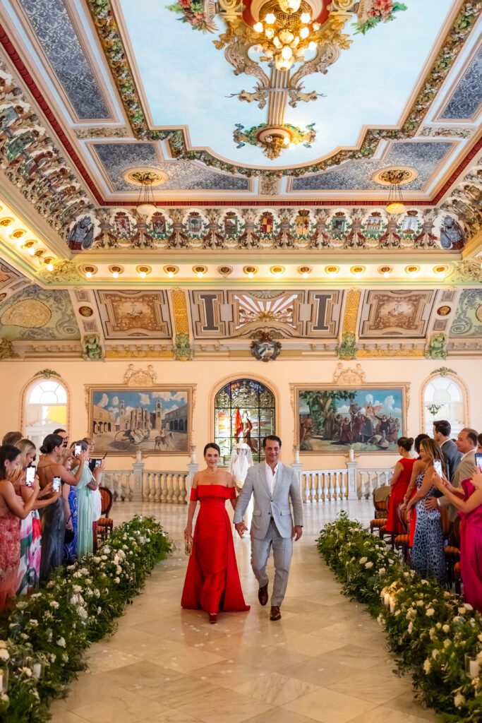 Los padres de Victoria avanzan hacia el altar mientras ella se detiene al inicio del camino en el Palacio de los Matrimonios de Prado.