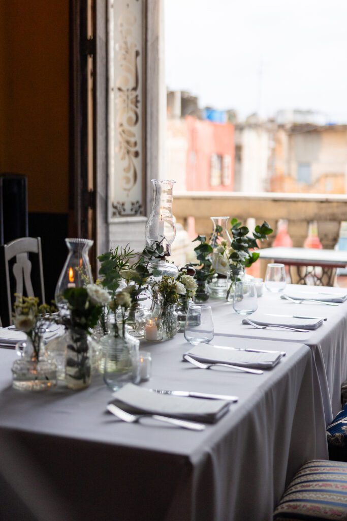 Mesa decorada con quinqués antiguos, flores blancas y greenery, con vista a las edificaciones de La Habana desde La Guarida