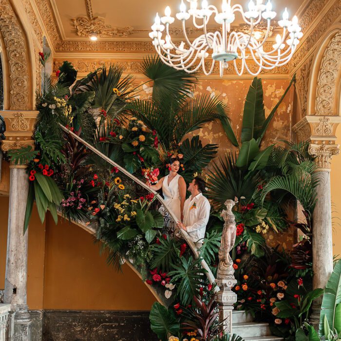 Los novios posando en la escalera de La Guarida, rodeados de flores tropicales, después de su boda.