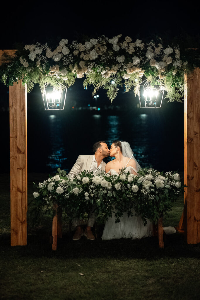 Novios se besan sentados bajo gazebo de flores blancas iluminado con farolas