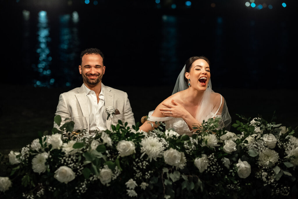 Novios ríen en la mesa presidencial rodeados de flores blancas