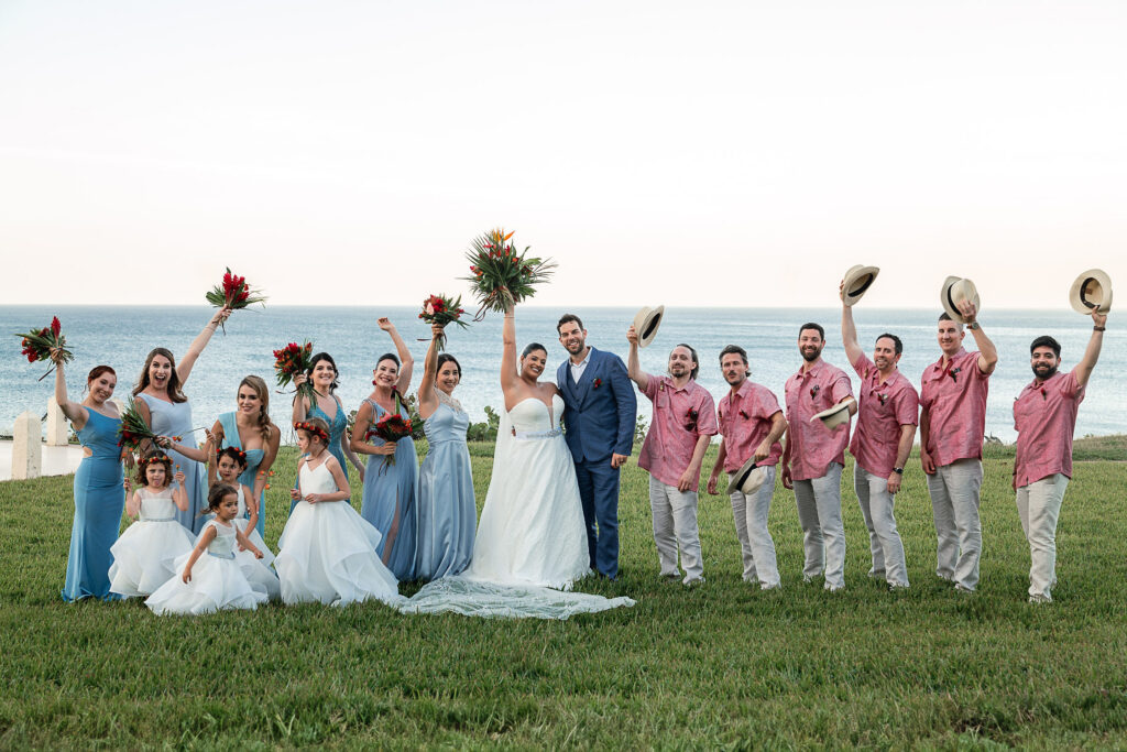 Couple with their wedding party and flower girls raising hats and bouquets by the sea at the Hotel Nacional