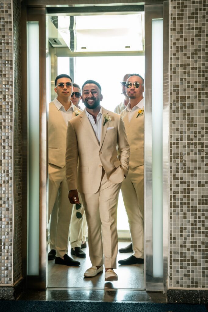 As the elevator doors open, the groom and his groomsmen walk in smiling