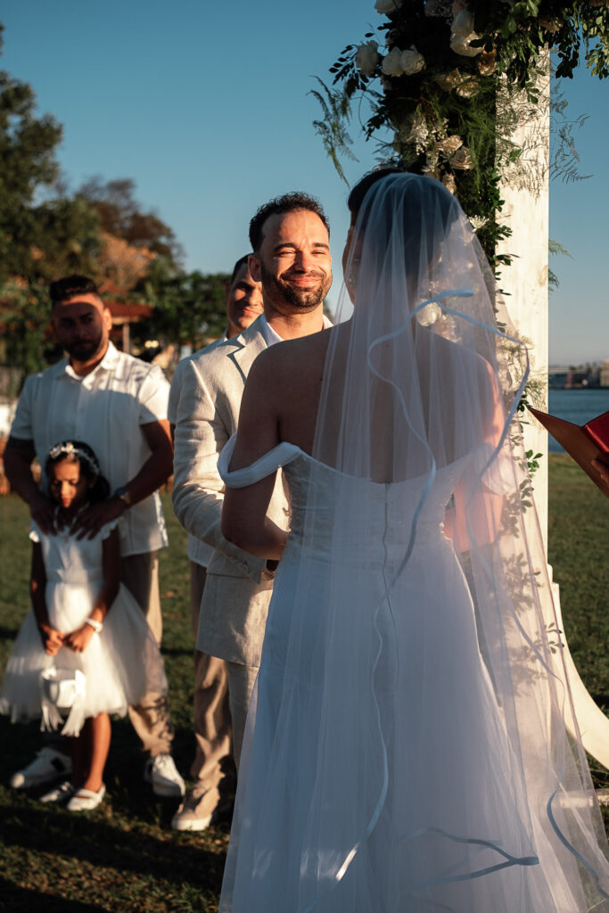 El novio observa a la novia con su corte al fondo y flores blancas decorando el gazebo