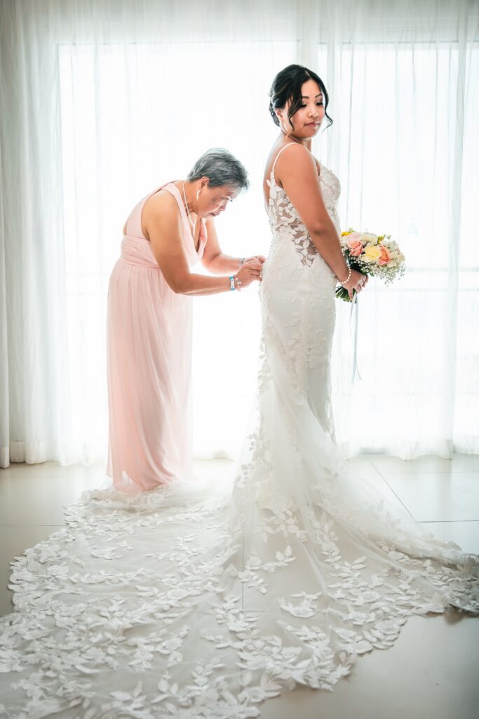 The mother adjusts the bride's dress by a sunlit window