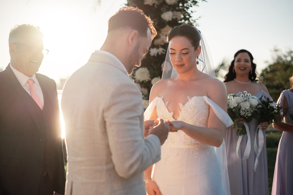El novio coloca el anillo a la novia con el sol y flores blancas detrás