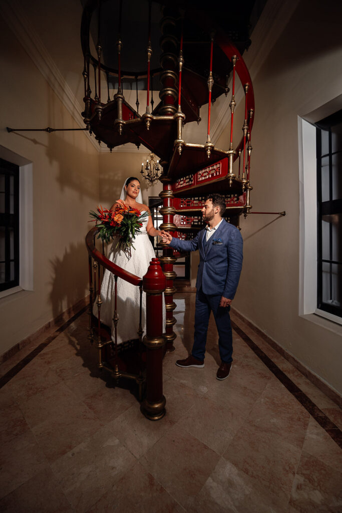 The groom helps the bride down a spiral wooden staircase with golden accents at the Hotel Nacional