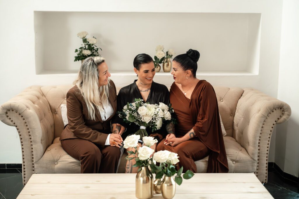 One of the brides sitting between both mothers, sharing laughs before getting dressed for the wedding