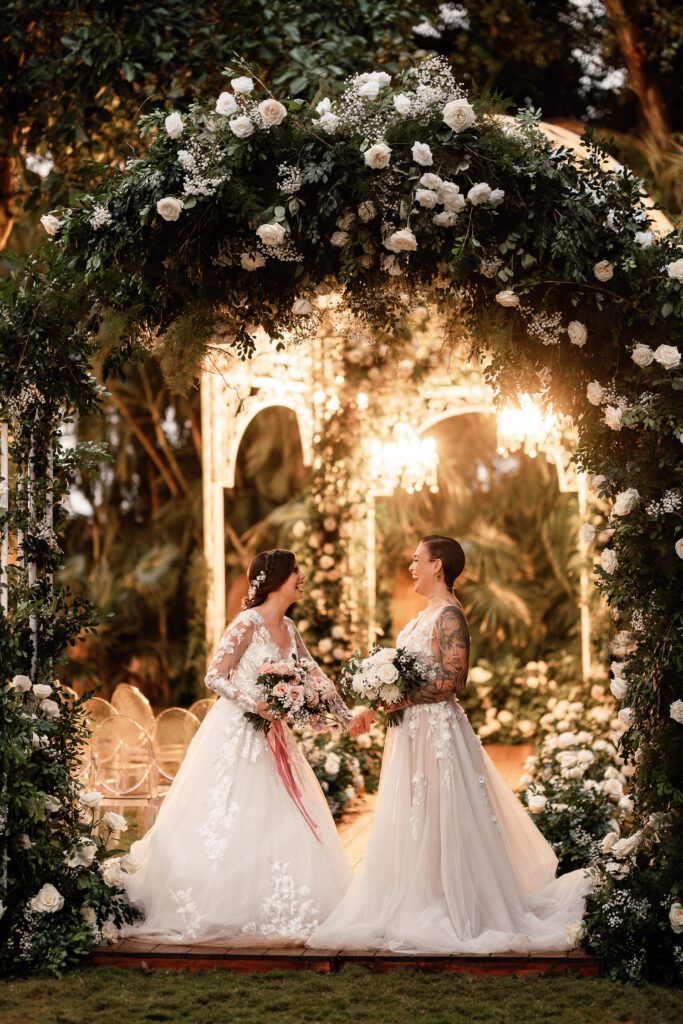 The brides smile under a floral arch with the dome lit up in the background at their wedding in Cuba under the most beautiful dome.
