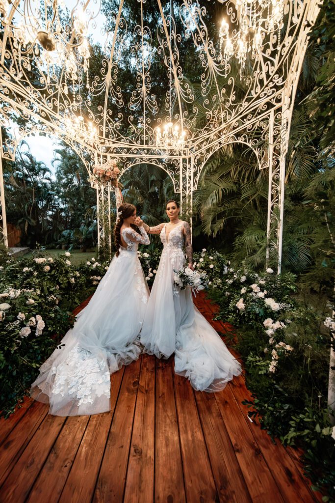 The brides pose under a dome decorated with flowers and lamps at their wedding in Cuba under the most beautiful dome