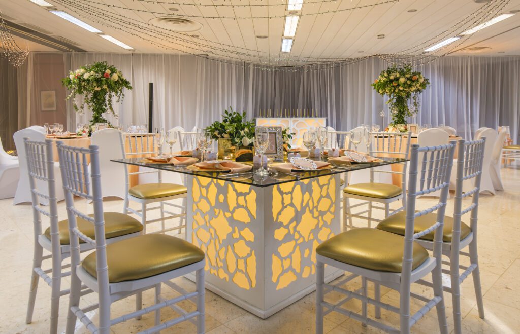 Vista de mesa cuadrada iluminada con sillas blancas y centros de mesa con follaje
