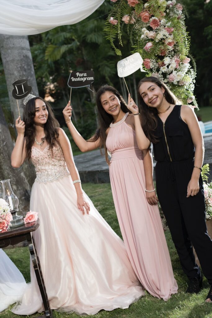 En una de nuestras fiestas de quince la quinceañera y dos amigas sonrientes posando con letreros divertidos en un photocall floral al aire libre