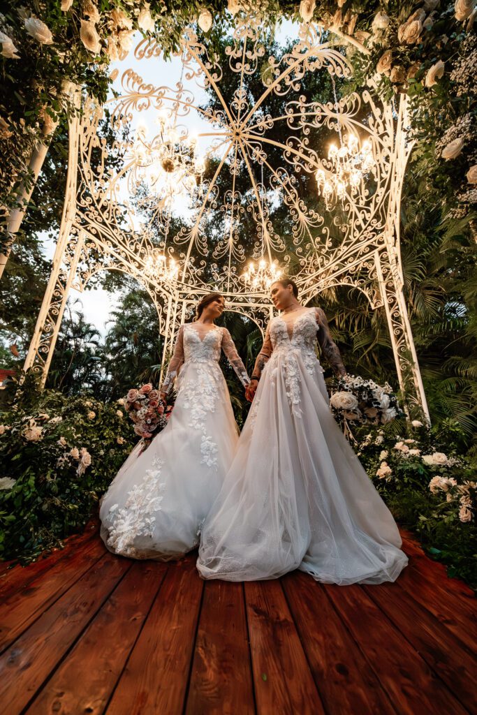 The brides smile at each other under an illuminated dome, surrounded by flowers at their wedding in Cuba under the most beautiful dome
