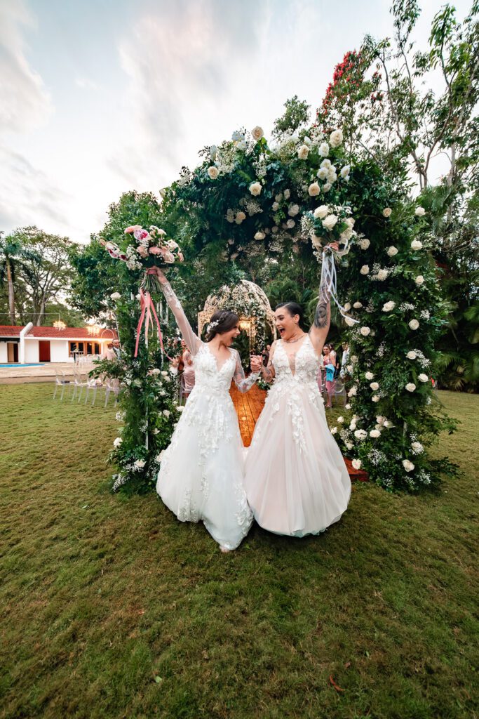 Two brides celebrate under a floral arch after their wedding ceremony in Cuba under the most beautiful dome